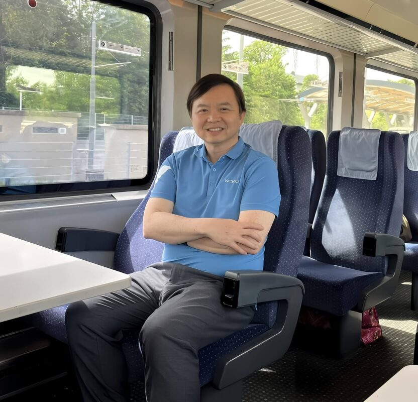專業導遊管延平穿著 KOMIA TORAY 藍色涼感快乾 POLO 衫與SUPPLEX抗 UV 內搭衣，坐在前往哈修塔特的維也納 OBB 火車窗邊。窗外是柔光的湖光山色，展現輕盈舒適的機能旅遊穿搭。